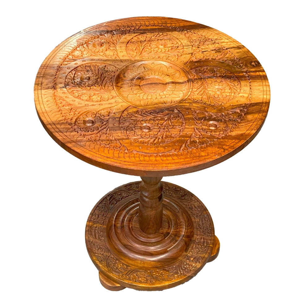 Handcrafted wooden pedestal table with intricate carved floral and paisley motifs covering the round tabletop and circular base, featuring a turned wooden pedestal stem with traditional carved details