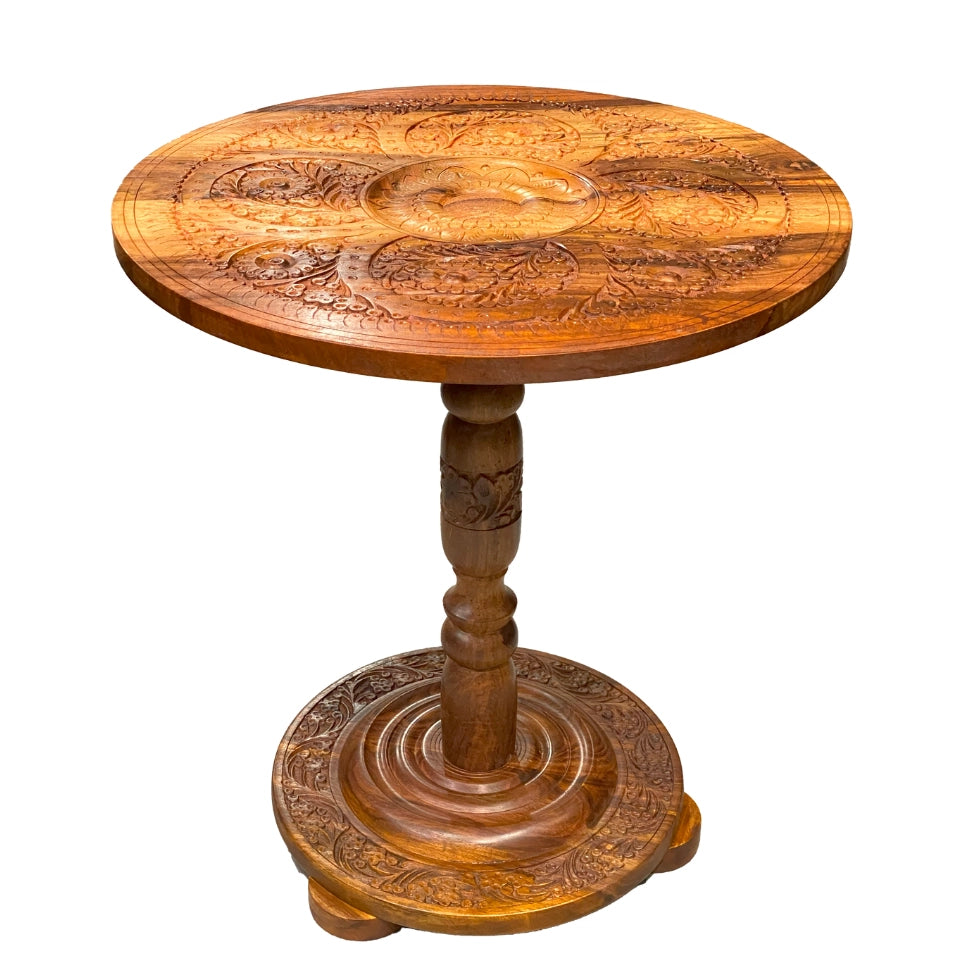 Handcrafted wooden pedestal table with intricate carved floral and paisley motifs covering the round tabletop and circular base, featuring a turned wooden pedestal stem with traditional carved details
